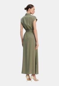 Vestito lungo verde oliva con maniche corte e vita cinturata. Presenta uno spacco laterale e una texture liscia, abbinato a scarpe con tacco alto.