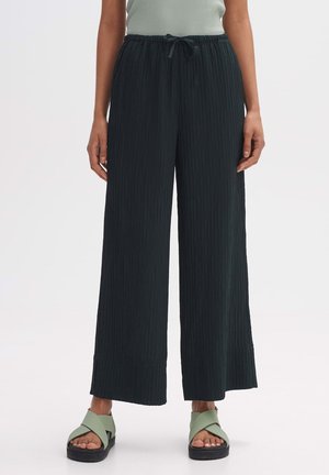 Wide-leg, dark green pants with textured fabric and a drawstring waist. Worn with light green sandals featuring a thick sole and crossed straps.