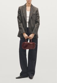 Woven burgundy handbag with a rounded shape, short handle, and zip closure, held by a person wearing a gray blazer and dark jeans.