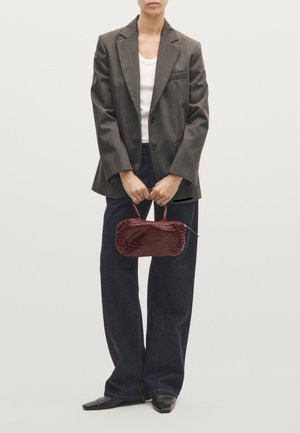 Gewebte burgunderfarbene Handtasche mit runder Form, kurzer Grifft und Reißverschluss, gehalten von einer Person in einem grauen Blazer und dunkler Jeans.