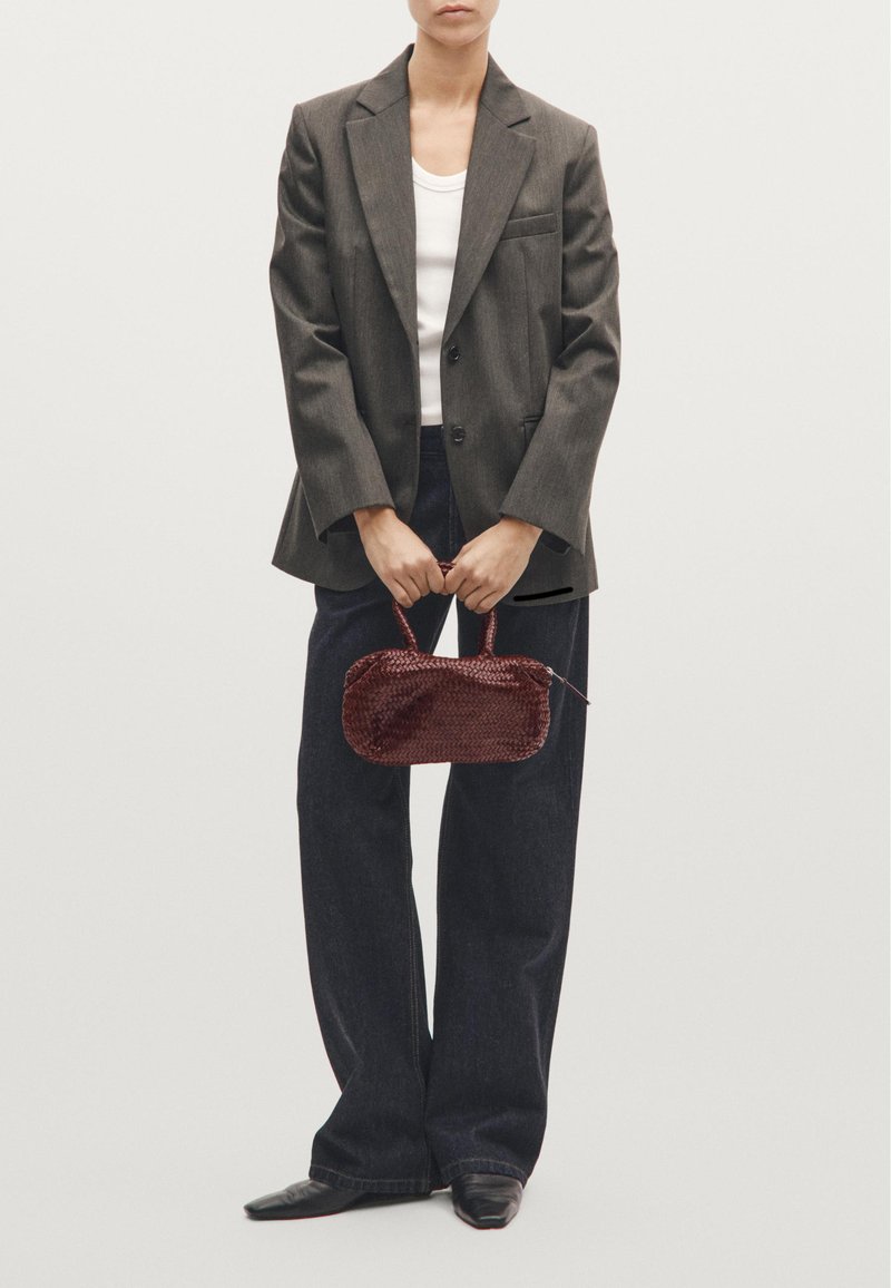 Vævet burgunder håndtaske med rund form, kort håndtag og lynlås, holdt af en person iført en grå blazer og mørke jeans.