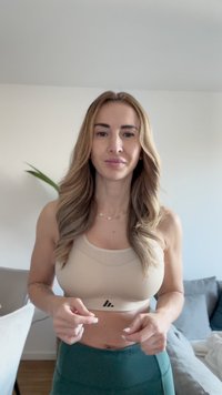 Woman with long wavy hair wearing a beige sports bra and green leggings, standing indoors with a plant and couch in the background.