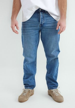 Muž v modrých džínsoch, bielom tričku a béžových teniskách stojí na jednoduchom bielom pozadí.