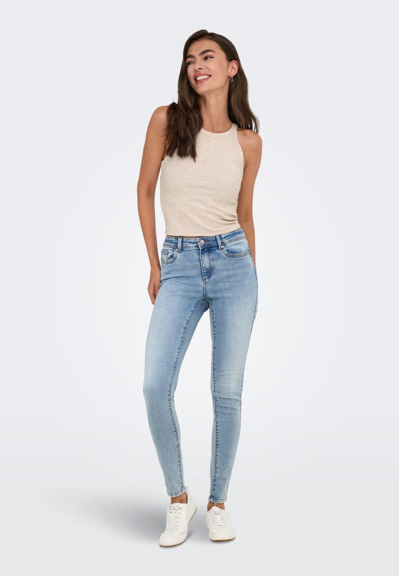 Femme aux longs cheveux bruns portant un haut beige sans manches, un jean skinny bleu clair et des baskets blanches, debout et souriante, les mains dans le dos.