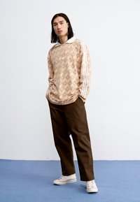 Long-sleeve beige shirt with geometric pattern, white collar, and three white stripes on sleeves; paired with loose brown trousers and white sneakers.