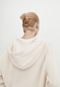 Crème-kleurige gebreide trui met een capuchon, met een gestructureerde stof, ribbelranden en een ontspannen pasvorm. Achteraanzicht toont haarstijl.