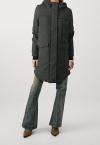 Mörkgrön lång parka med huva, två framfickor och ribbade ärmslut. Bärs över utsvängda blå jeans och bruna klackade ankelskor.