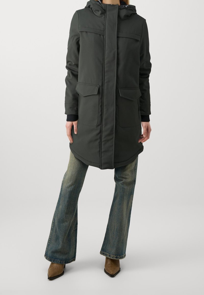 Mörkgrön lång parka med huva, två framfickor och ribbade ärmslut. Bärs över utsvängda blå jeans och bruna klackade ankelskor.