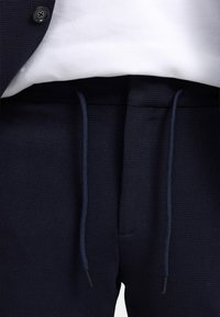 Granatowe joggery z teksturowanej tkaniny z elastycznym paskiem w talii, posiadające ciemnoniebieskie sznureczki i kieszeń z przodu. Uzupełnione białą koszulą na górze.