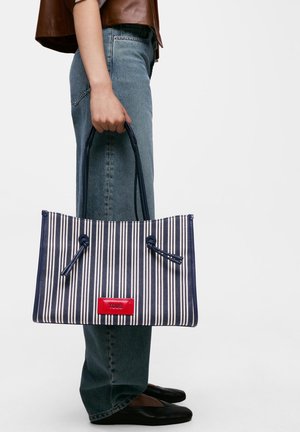 Persona que lleva vaqueros azules y camiseta marrón sosteniendo un bolso tote a rayas azul marino y blanco con etiqueta roja "TOUS", de pie frente a fondo blanco.