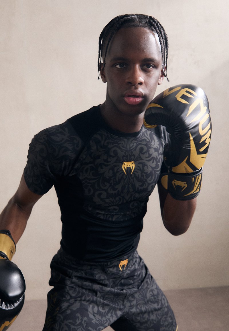 Camisa atlética negra con un patrón intrincado; pantalones cortos a juego. Guantes amarillos con el nombre de la marca. Enfoque en la pose, mostrando disposición para el boxeo.