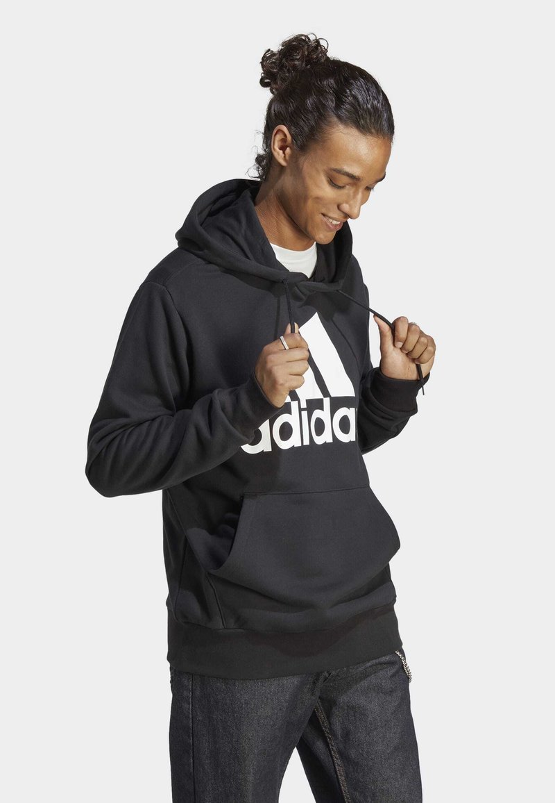 Hoodie noir avec poche kangourou, capuche à cordon de serrage et grand logo Adidas blanc sur le devant. Texture lisse, coupe décontractée et manches longues.