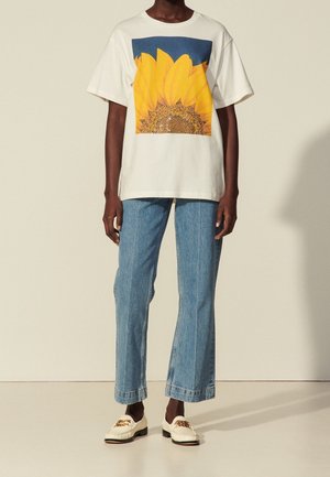 Person trägt weißes T-Shirt mit großem Sonnenblumendruck, hellblaue Jeans mit geradem Bein und weiße Loafer mit Kettendetail.