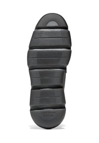 Svart gummisul med texturerade mönster och en rundad klack. Sulan har flera räfflor och en detalj med "Cole Haan Grand 360"-logotyp.