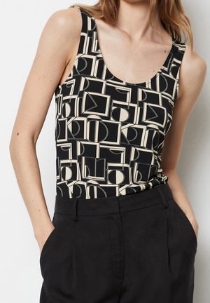 Frau trägt ein ärmelloses schwarz-beiges Top mit geometrischem Muster, in hoch taillierte schwarze Hosen gesteckt, mit den Händen in den Taschen.