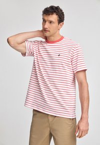 Röd och vit randig t-shirt med kort ärm och rund halsringning, med en logotyp på bröstet, tillsammans med khakibyxor.