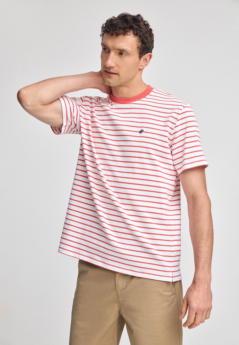 Röd och vit randig t-shirt med kort ärm och rund halsringning, med en logotyp på bröstet, tillsammans med khakibyxor.