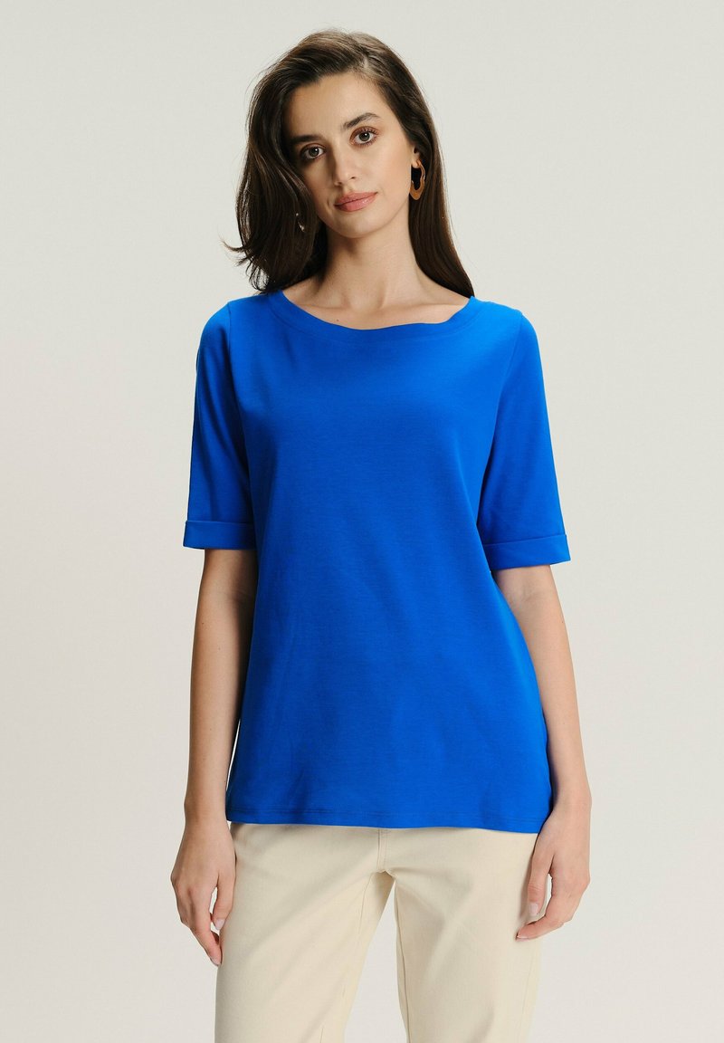 Kobieta o długich ciemnych włosach, ubrana w krótką niebieską koszulę i beżowe spodnie, stojąca na jednolitym jasnym tle.