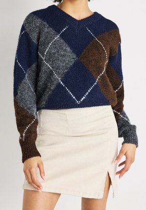 Person iført en navyblå, brun og grå argyle strikket sweater kombineret med en beige kordnøgle mini nederdel med frontslids.
