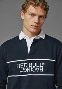 Námořnická modrá polo košile s dlouhým rukávem, bílým límcem a logem Red Bull Racing. Obsahuje bílé horizontální pruhy na hrudi. Bavlněný materiál.