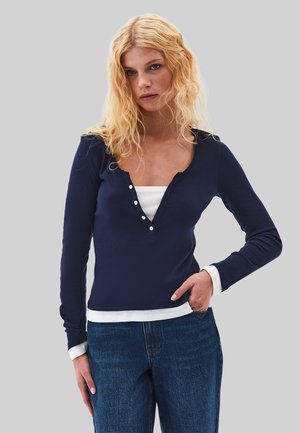 Young woman with blonde curly hair wearing a navy long-sleeve top over a white undershirt and blue jeans, standing against a gray background.
