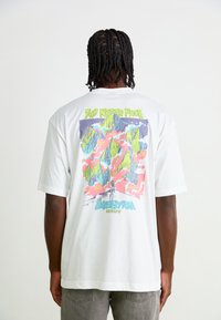 Hvid t-shirt med et farverigt bjergmotiv på ryggen, med grønne, blå og lyserøde detaljer, samt korte ærmer.