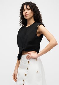 Μαύρο crop top με κόμπο στη μέση, διακοσμημένο με χρυσές λεπτομέρειες στον ώμο. Συνοδεύεται από λευκή φούστα ψηλόμεση με χρυσά κουμπιά.