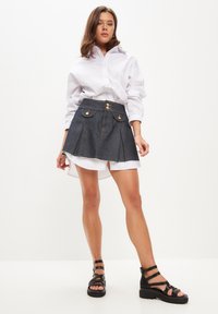 Denim mini suknja s plješcima, poderanim rubom, zlatnim dijelovima i dvije prednje džepove. Uparena s bijelom oversized košuljom na gumb i crnim sandalama.
