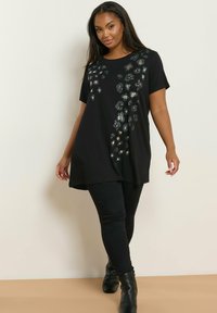 Nainen pukeutuneena mustalla kukkakuosisella tunikalla, mustilla slim-fit-housuilla ja mustilla nilkkureilla, seisoo yksinkertaista vaaleaa seinää vasten.