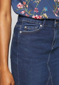 Džínová sukně v tmavě modré barvě, s upnutým střihem, která má malou přední kapsu a ozdobné prošívání. Texturovaná tkanina.