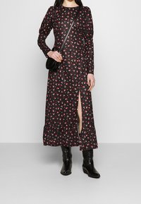 Kvinna klädd i en svart långärmad blommig klänning med slits framtill, svart crossbody-väskan och svarta läderstövlar till mitten av vaden som står på en enfärgad bakgrund.