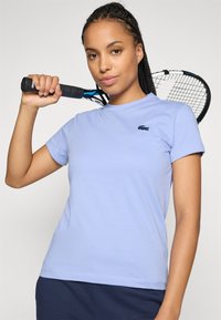 Femeie cu părul împletit, îmbrăcată cu o cămașă albastră deschisă, ține o rachetă de tenis deasupra umărului și se uită cu încredere la cameră.