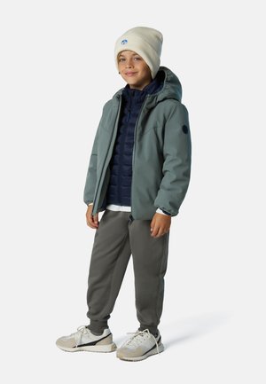 Niño con una chaqueta impermeable verde claro sobre un chaleco acolchado azul marino, pantalones deportivos grises y zapatillas beige con detalles y cordones blancos.
