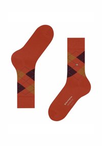 Een paar gebrand-oranje sokken met een argylepatroon in donkerrood, mosterdgeel en bruin, met kleine merktekst en een metalen embleem op één van de sokken.