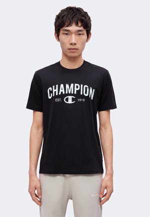 Νέος άνδρας που φοράει μαύρο μπλουζάκι Champion με λευκό λογότυπο και ανοιχτογκρι παντελόνι σε απλό φωτεινό φόντο.