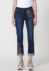 Temno modri jeans s edinstvenimi večbarvnimi vezeninami na sprednji strani. Ima zvite manšete in klasičen dizajn s петimi žepi.