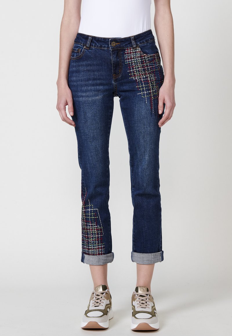 Temno modri jeans s edinstvenimi večbarvnimi vezeninami na sprednji strani. Ima zvite manšete in klasičen dizajn s петimi žepi.