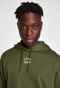 Bluza z kapturem w kolorze oliwkowej zieleni, wykonana z miękkiego materiału, wyposażona w sznurek do regulacji kaptura, biały logo z przodu i luźny krój.