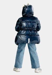 Mörkblå puffjacka med en glänsande yta, insnörd midja och huva. Kombinerad med ljusblå vida jeans och vita sneakers.