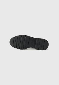 Suela de zapato de goma negra con surcos y crestas texturizadas, que presenta una impresión de logo y un diseño de punta redondeada.