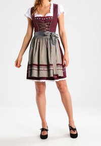 Kobieta w tradycyjnej bordowo-beżowej sukni dirndl z białą bluzką i czarnymi balerinkami stoi na tle białej ściany.