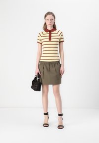 Stripet poloshirt i gul og maroon, paret med en olivengrønn skjørt med lommedetaljer, og svarte helsandesko. Holder en svart veske.