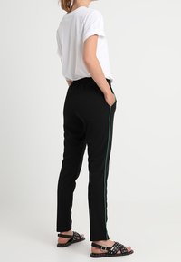 Svarta byxor med smal passform och grön sidostrimma, kombinerade med en vit t-shirt med rullade ärmar och svarta sandaler med nitar.