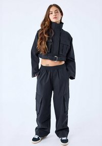 Zwarte cropped jas met een hoge kraag, voorkant met rits en meerdere zakken, gecombineerd met losse zwarte broek met zijzakken.