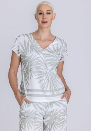 Vrouw met kort blond haar, gekleed in een wit shirt en een broek met lichtgrijze palmbladpatronen, handen in de zakken, neutrale achtergrond.