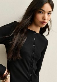 Czarny prążkowany top z długim rękawem z guzikowym zapięciem z przodu, okrągłym dekoltem i gładką fakturą. Podkreślony błyszczącymi czarnymi guzikami.