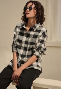 Černobílé kostkované flanelové tričko s volným střihem a zapínáním na knoflíky, v kombinaci s černými džínami a kulatými slunečními brýlemi.