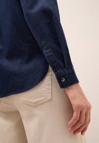 Mörk denimskjorta med knappmanchett, med en lätt rundad nederkant. Beiga byxor med sydd detalj på bakfickan. Slät textur överlag.