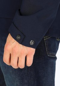 Marineblå jakke i stof med en glat tekstur; har en knapmanchet med mat sort hardware; denim jeans synlige med en falmet finish.