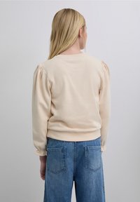 Hanorac bej din țesătură moale, cu mâneci bufante și manșete rib, asortat cu pantaloni largi din denim albastru, prevăzuți cu două buzunare pe spate.